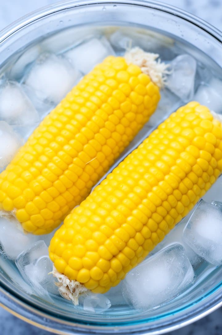 Overhead shot of blanched corn ears resting in a large glass bowl filled with ice water and ice cubes, the corn bright yellow against the clear water and white ice, cool blue-white tones, natural daylight from above, droplets of water on the corn silk remnants