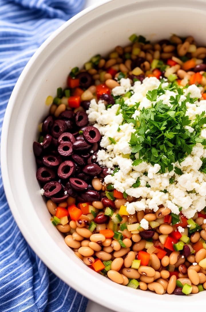 Large white mixing bowl filled with all the salad components before dressing — beans and diced vegetables layered with sliced kalamata olives, crumbled feta, and chopped parsley on top, showing the vibrant color contrast, overhead angle, natural light, blue striped towel beside the bowl