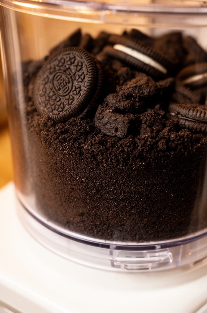 Close-up shot of Oreo cookies being crushed in a food processor, half-processed with fine dark crumbs at the bottom and larger cookie chunks at the top, clear processor bowl allowing view of the texture gradient, shot from 45-degree angle, warm kitchen lighting, shallow depth of field focusing on the crumb texture, white countertop below