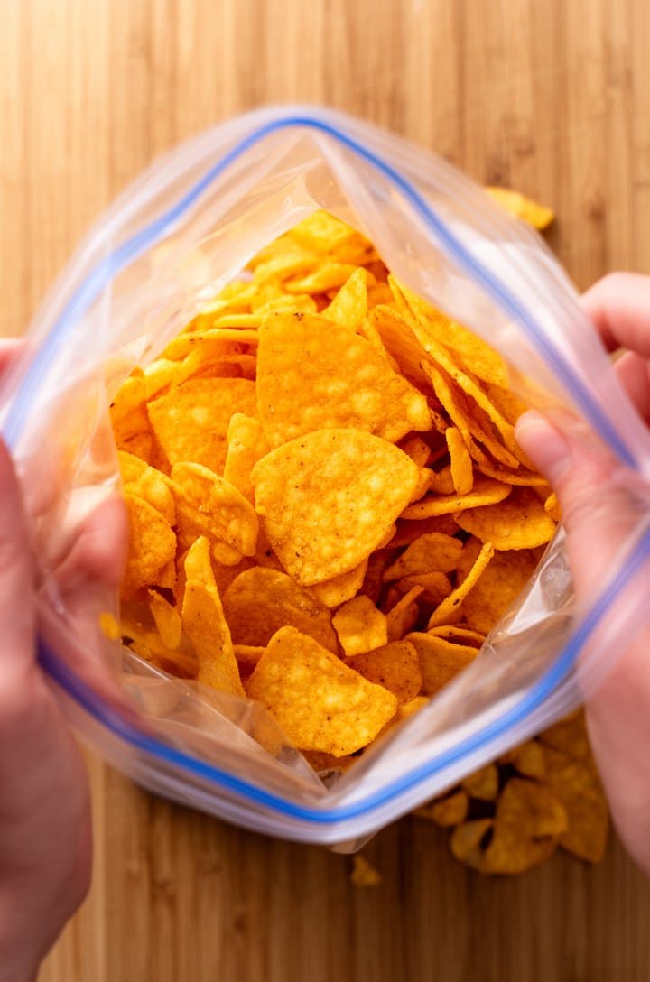 Top-down overhead shot of hands crushing Frito corn chips inside a clear zip-lock bag on a wooden cutting board, golden-orange chip pieces in various sizes from large shards to small crumbles, natural lighting, casual kitchen scene