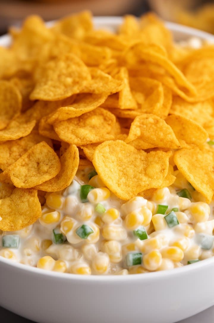 Extreme close-up of the finished Frito corn salad in a white ceramic serving bowl, crushed golden Fritos layered generously over the top of the creamy corn mixture, individual corn kernels and tiny green pepper dice visible beneath the chips, shallow depth of field, warm natural side lighting highlighting the crispy chip textures and creamy dressing