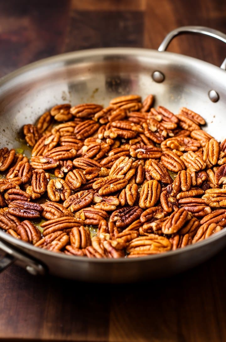 Close-up of golden toasted pecan halves in a dry stainless steel skillet, some pieces slightly darker with visible oil sheen from toasting, warm amber tones, shot from 45-degree angle with shallow depth of field, dark wooden cutting board visible in the background.