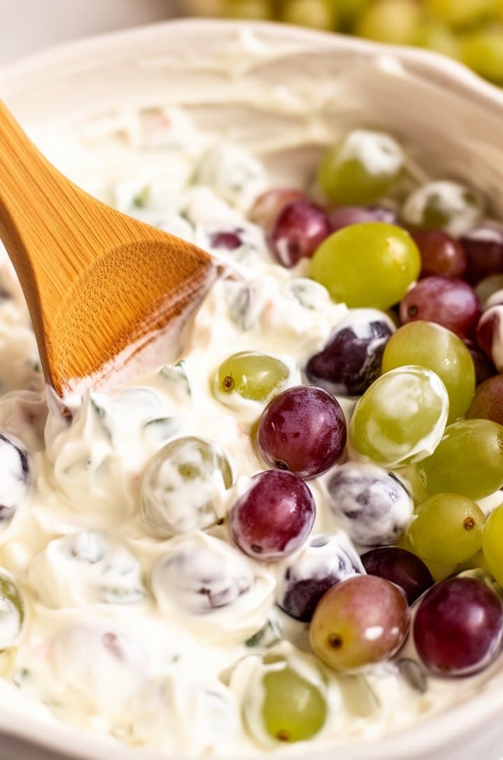 Side-angle close-up of red and green grapes being folded into the white cream cheese dressing with a large wooden spoon, half the grapes coated and glistening, the other half still showing bare purple and green skins, bright kitchen lighting, creamy bokeh background.