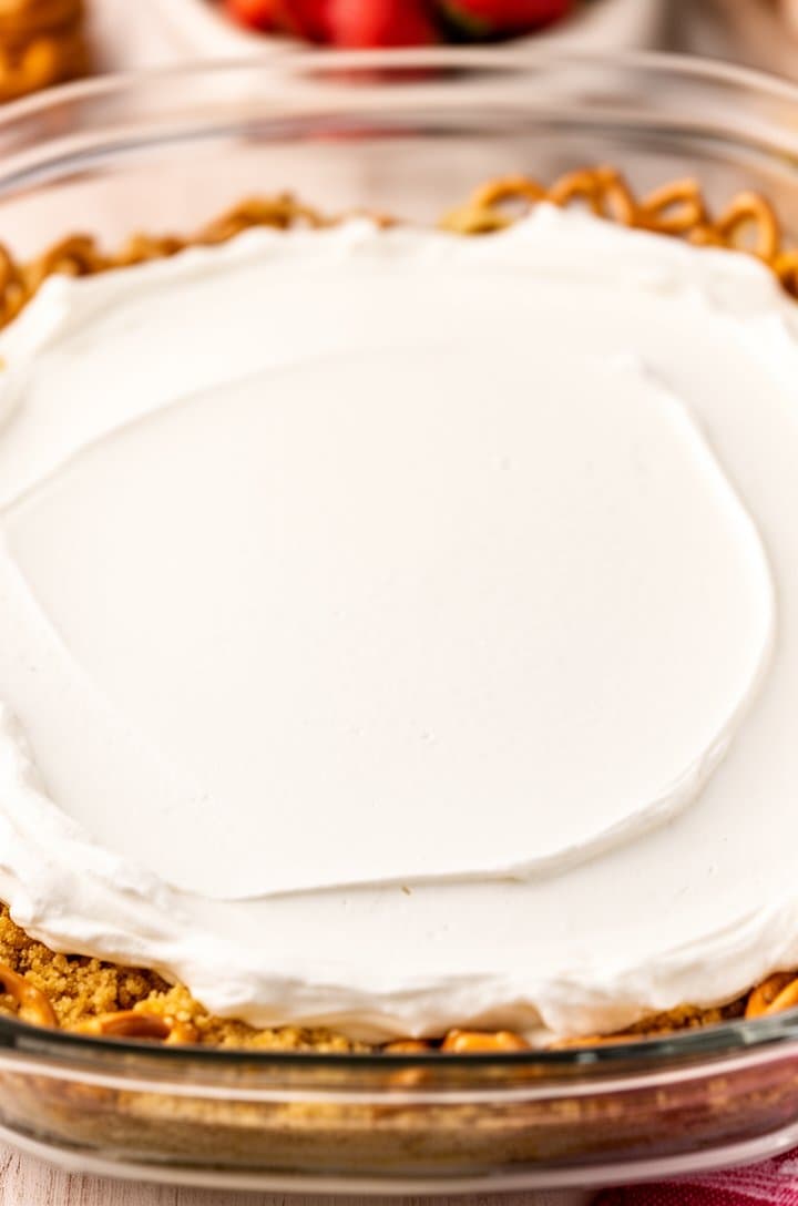Overhead shot of the cream cheese layer fully spread over the pretzel crust in the glass baking dish, perfectly smooth and white like fresh snow, sealed completely to all four edges with no gaps visible, the golden pretzel crust peeking through slightly at the very edges, bright even lighting, clean composition