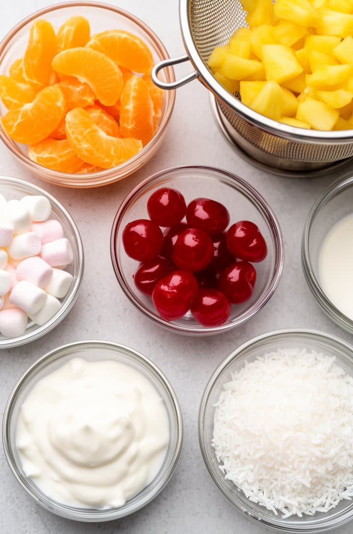 Overhead flat-lay of all ambrosia ingredients in individual small glass prep bowls arranged on a light gray surface — mandarin oranges, pineapple tidbits, maraschino cherries, mini marshmallows, shredded coconut, Greek yogurt, and heavy cream. A colander with drained fruit nearby. Bright even natural lighting from above, clean organized composition, professional food blog photography