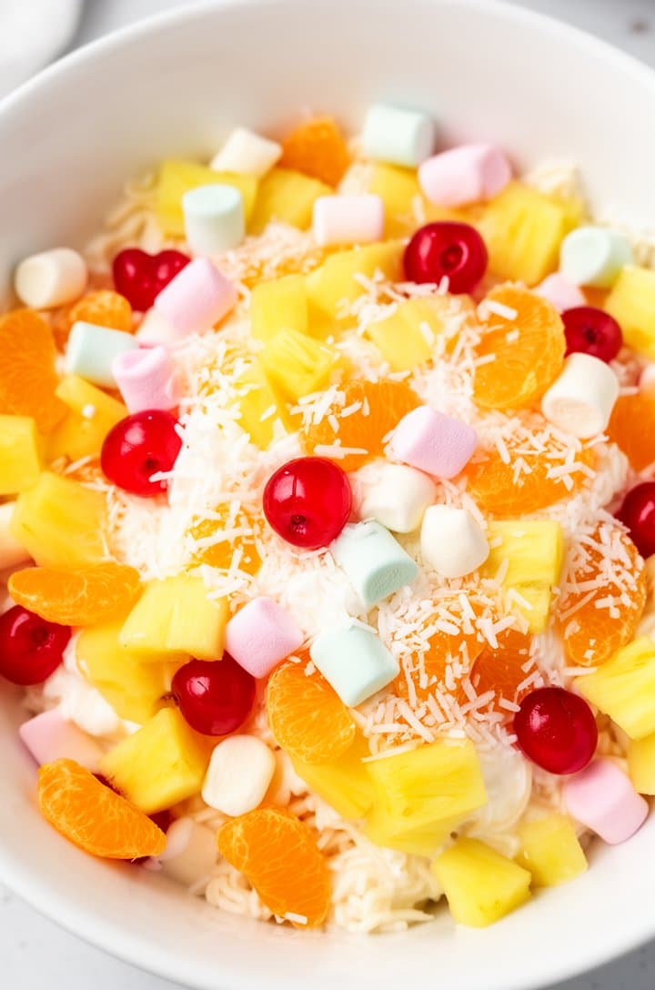 Overhead shot of colorful fruit mixture — orange mandarin segments, yellow pineapple chunks, bright red cherry halves, pastel mini marshmallows, and white shredded coconut — in a large white bowl before the cream is added. Vibrant colors against the white bowl, bright natural lighting, sharp focus across the entire bowl