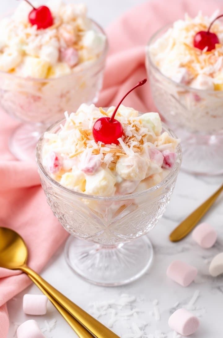 Finished ambrosia fruit salad served in individual crystal cut-glass dessert bowls on a white marble surface, garnished with extra coconut shreds and a single maraschino cherry on top of each. Gold dessert spoons alongside, a soft pink linen napkin draped between the glasses, scattered coconut and marshmallows on the surface. Bright airy natural lighting, pastel color palette, styled from a 30-degree overhead angle