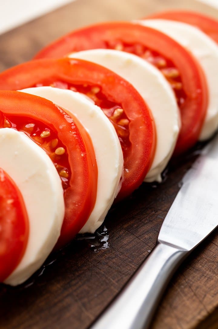 Close-up shot of thick 1/4-inch tomato rounds and matching mozzarella slices arranged side by side on a dark wooden cutting board, a sharp knife resting alongside, tomato juice glistening on the cut surfaces, bright natural side lighting highlighting the texture of each slice, shallow depth of field