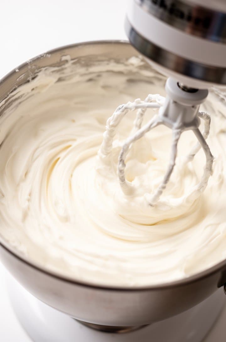 Medium shot of cream cheese frosting being beaten in a stand mixer bowl, showing the white fluffy mixture with visible paddle attachment tracks creating smooth swoops, the frosting looks thick and silky with a glossy sheen, bright overhead lighting, clean white kitchen background