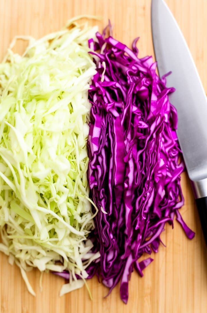 Overhead flat-lay of finely shredded napa cabbage and red cabbage side by side on a light wood cutting board, showing the contrast between the pale green-white napa and deep purple-magenta red cabbage, a sharp chef's knife resting to the side, bright even natural daylight from above, clean minimalist food prep photography