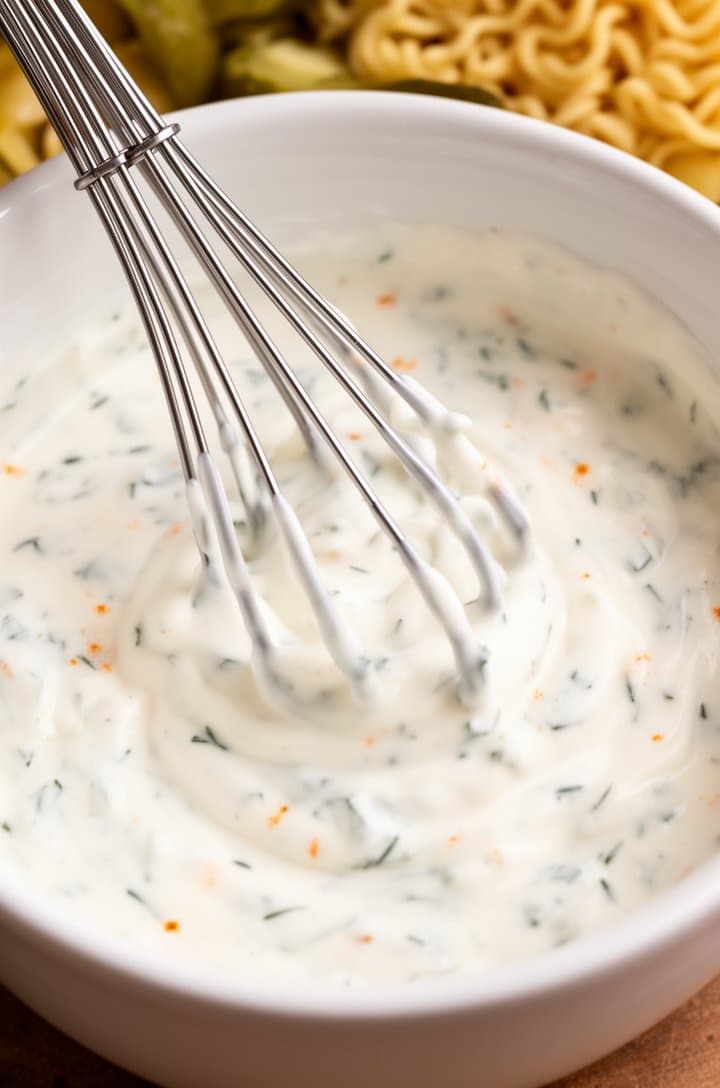 Overhead close-up of a small white bowl containing the creamy dressing being whisked with a small wire whisk — thick white mixture of mayonnaise and sour cream with visible tiny green dill flecks and a hint of orange from cayenne, smooth and glossy texture, bright even lighting