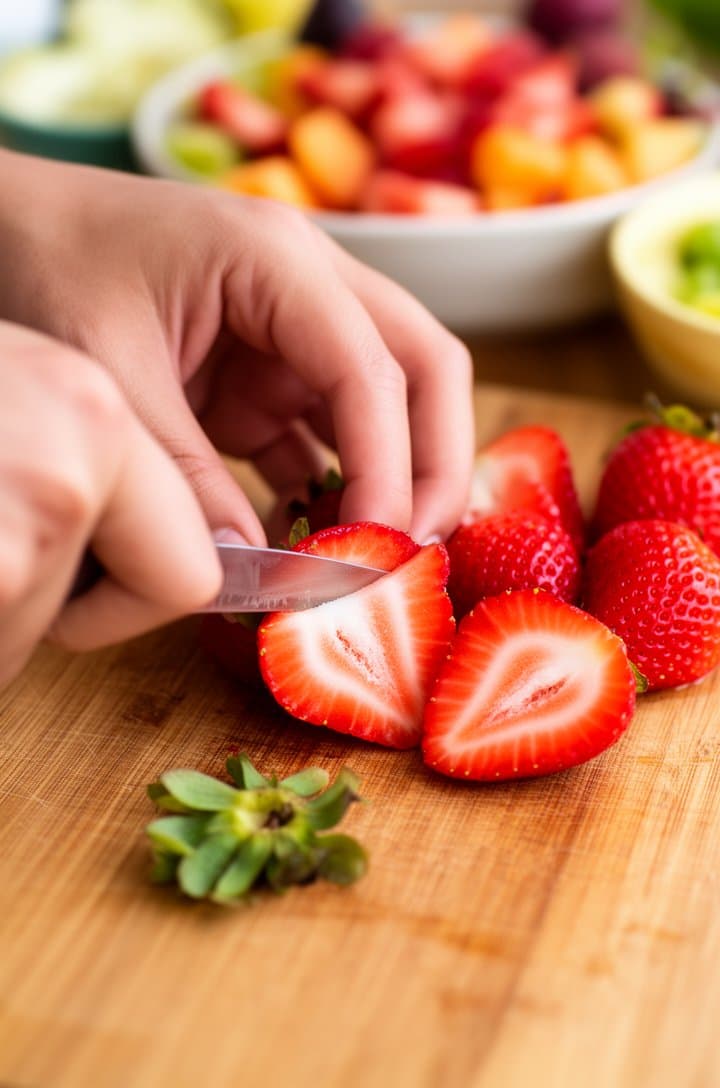 Close-up of hands slicing fresh strawberries on a wooden cutting board, showing the bright red interior and white center of each berry, a small pile of hulled stems to the side, soft natural lighting from the left, shallow depth of field with other prepped fruits blurred in bowls behind
