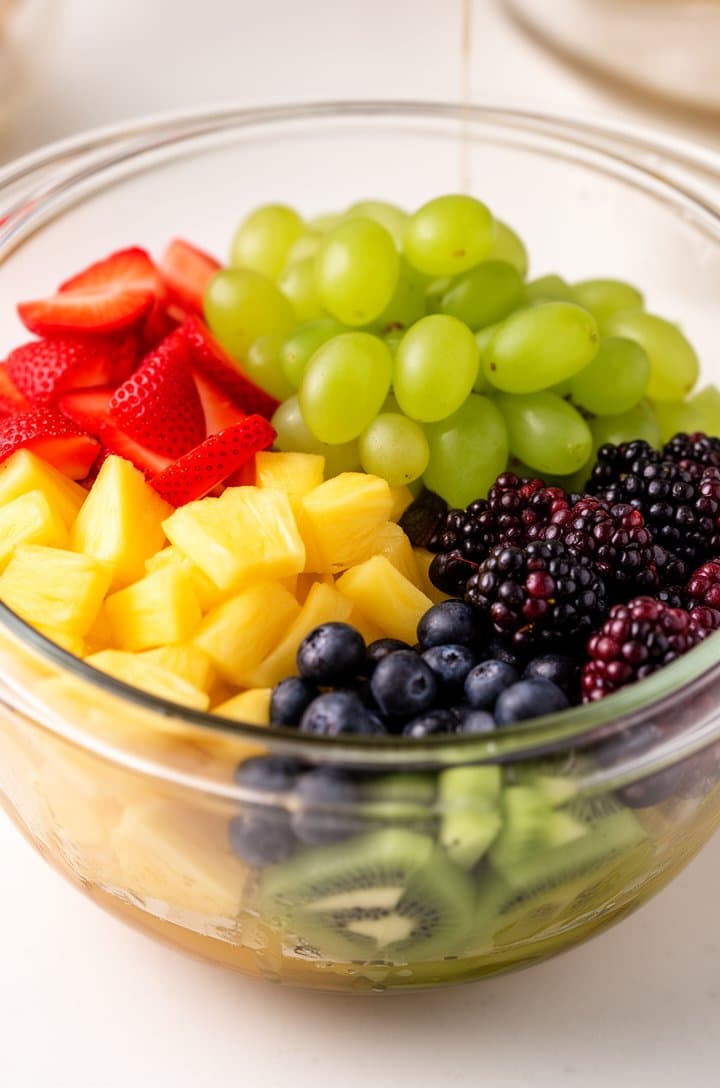 45-degree angle shot of all chopped fruits being added to a large clear glass mixing bowl — layers of red strawberry slices, green grapes, golden pineapple, dark blueberries, purple blackberries, and green kiwi visible through the glass sides, bright natural lighting, white countertop surface