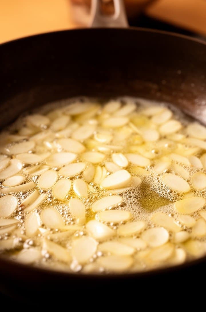 Close-up angled shot of thinly sliced garlic pieces gently cooking in foaming melted butter in a large dark skillet, each slice turning light golden at the edges, tiny bubbles surrounding the garlic, warm amber-gold tones throughout, shallow depth of field with the skillet handle blurred in the background, warm side lighting