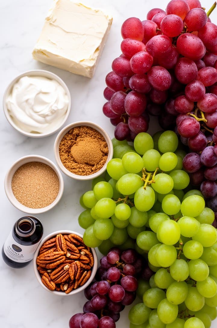 Overhead flat-lay of all grape salad ingredients neatly arranged on a white marble surface — a block of softened cream cheese, small bowl of sour cream, ramekins of granulated sugar and brown sugar, vanilla extract bottle, clusters of red and green seedless grapes freshly washed with water droplets, small bowl of raw pecans. Bright, even natural lighting from above, clean styling with minimal props