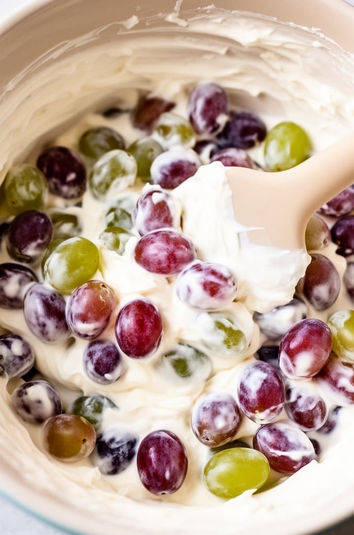 Overhead shot of red and green grapes being gently folded into the white cream cheese dressing in a large mixing bowl, some grapes fully coated and others still showing their colorful skins, a silicone spatula mid-fold, the contrast of purple-red and green against white dressing, bright natural lighting