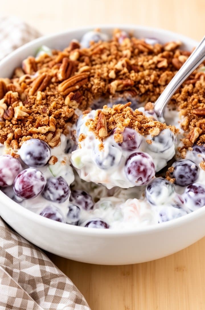 Finished grape salad served in a white ceramic bowl on a light wooden table, topped generously with brown sugar pecan crumble, a serving spoon scooping out a portion showing the creamy interior with mixed grape colors, a checkered linen napkin beside the bowl, bright natural daylight, inviting and appetizing composition