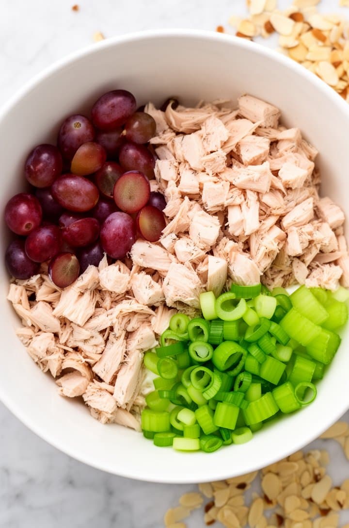 Overhead flat-lay of diced cooked chicken breast, halved seedless red grapes, diced celery stalks, and thinly sliced green onions arranged in a large white ceramic bowl before mixing, on a light marble surface with scattered toasted almond slivers nearby, bright even natural lighting from above, clean and appetizing