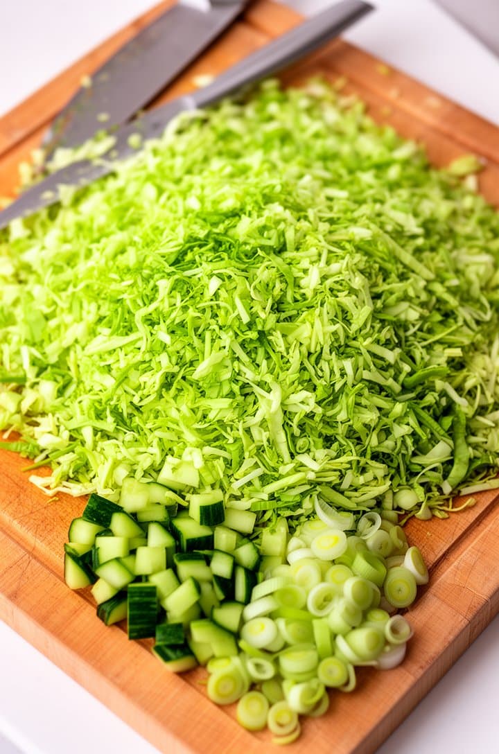 Close-up overhead shot of a large wooden cutting board covered with finely chopped confetti-sized green cabbage pieces, showing the tiny uniform size compared to a chef's knife blade resting at the edge. Small separate piles of diced cucumber and sliced green onion rounds nearby. Bright natural lighting from above, sharp focus on the vegetable textures, clean white countertop visible around the cutting board edges