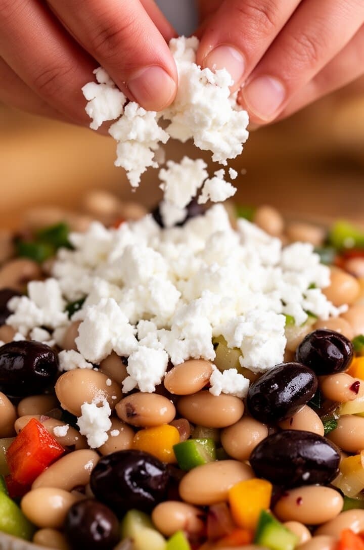 Close-up of white feta cheese being crumbled by hand over the top of the dressed bean salad, small irregular chunks falling onto the glistening beans and vegetables below. Fingers visible at the top of the frame, shot from slightly above, warm natural lighting, the colorful salad filling the lower two-thirds of the frame with beautiful contrast between white feta and dark olives