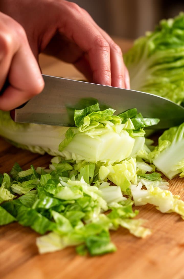 Close-up of hands chopping crisp green romaine lettuce on a wooden cutting board, the knife mid-slice through the pale green-white inner leaves, loose chopped pieces scattered on the board, whole romaine hearts visible to the side. Warm natural side lighting, shallow depth of field focusing on the blade and freshly cut lettuce, kitchen background softly blurred