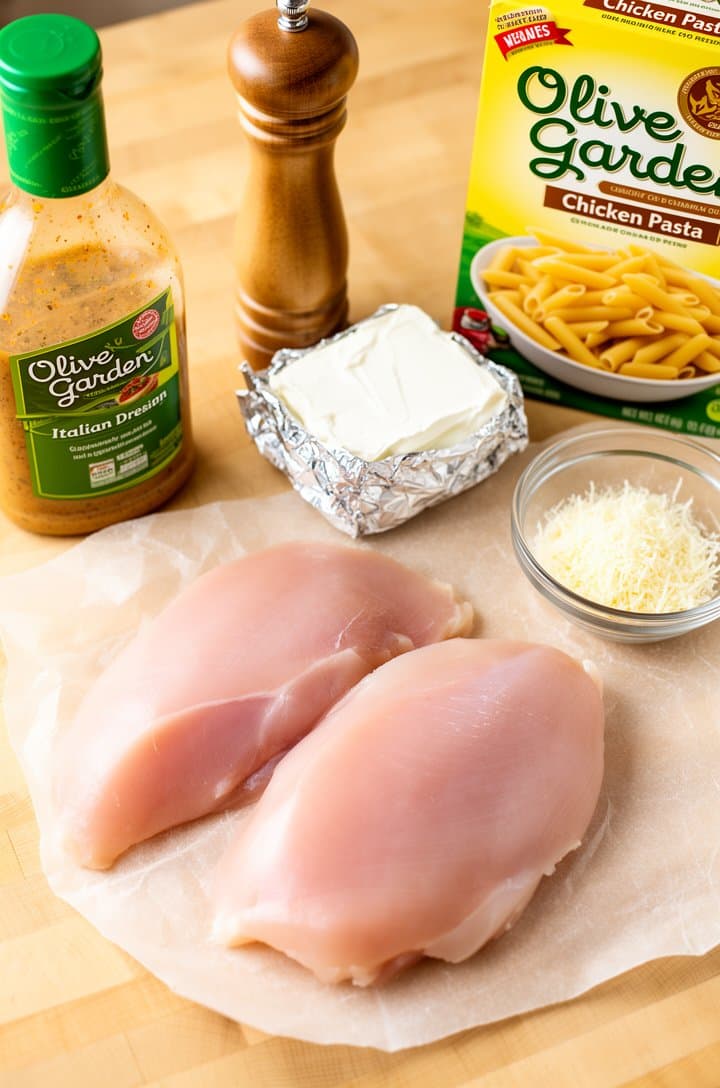 Overhead flat-lay of raw chicken breasts on parchment paper beside an open bottle of Olive Garden Italian dressing, a foil-wrapped block of cream cheese, a small glass bowl of grated parmesan, a box of penne pasta, and a pepper grinder, all arranged on a light butcher block countertop with bright natural window lighting and soft shadows