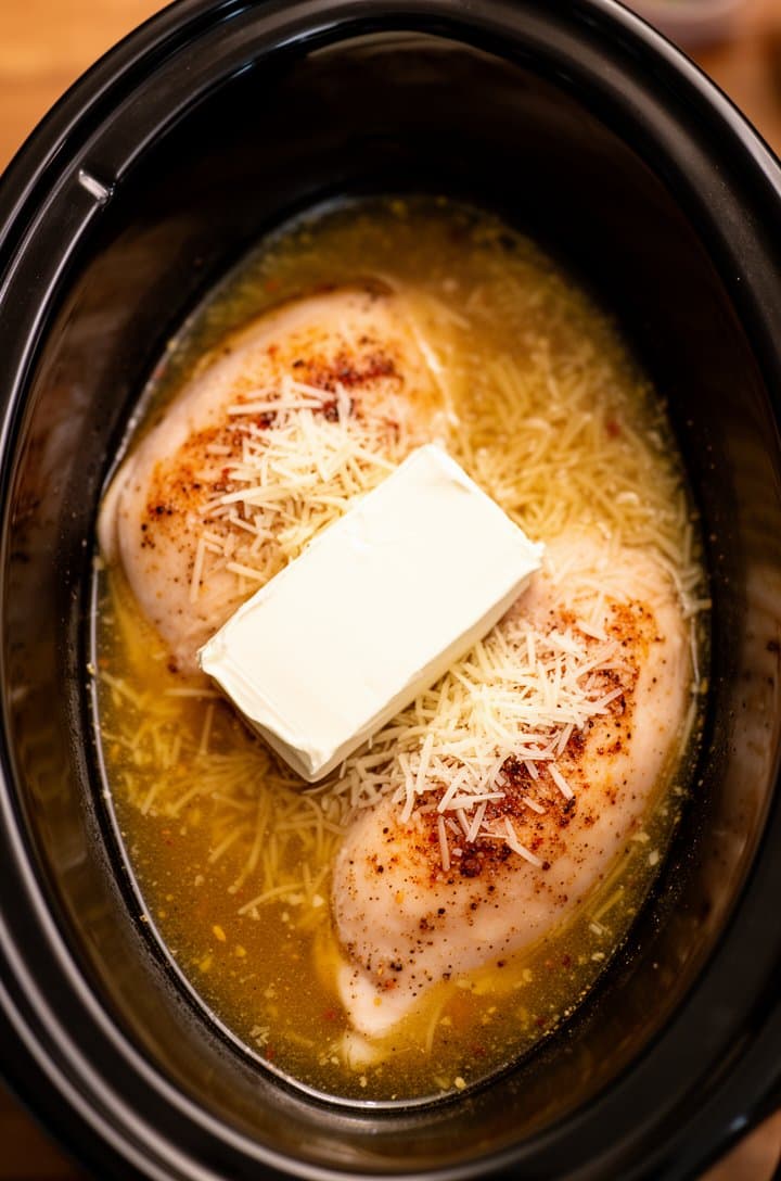 Straight-down overhead shot into a black oval slow cooker with two chicken breasts submerged in golden Italian dressing, a whole block of cream cheese placed on top of the chicken, parmesan cheese sprinkled over everything, before the lid goes on, warm kitchen lighting from the side