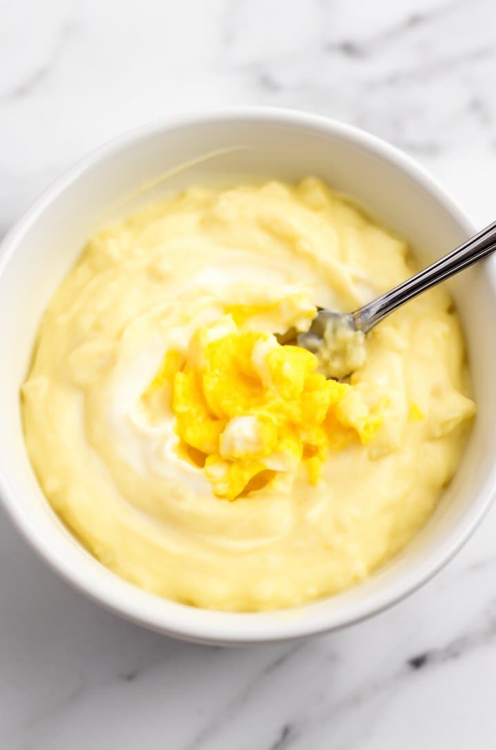 Overhead close-up of a small white bowl containing the dressing — mashed egg yolks blended with mayonnaise, yellow mustard, and buttermilk into a smooth creamy pale yellow mixture. A fork with remnants of mashed yolk resting in the bowl. Bright natural lighting, white marble surface, clean and appetizing