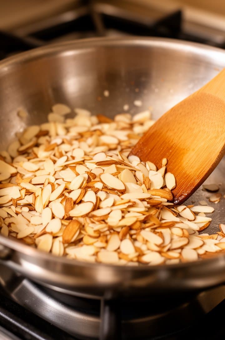 Close-up of a dry stainless steel skillet on a gas burner with slivered almonds being toasted, some pieces turned golden brown while others are still pale, a wooden spatula mid-stir. Warm overhead lighting, shallow depth of field focusing on the almonds in the center of the pan