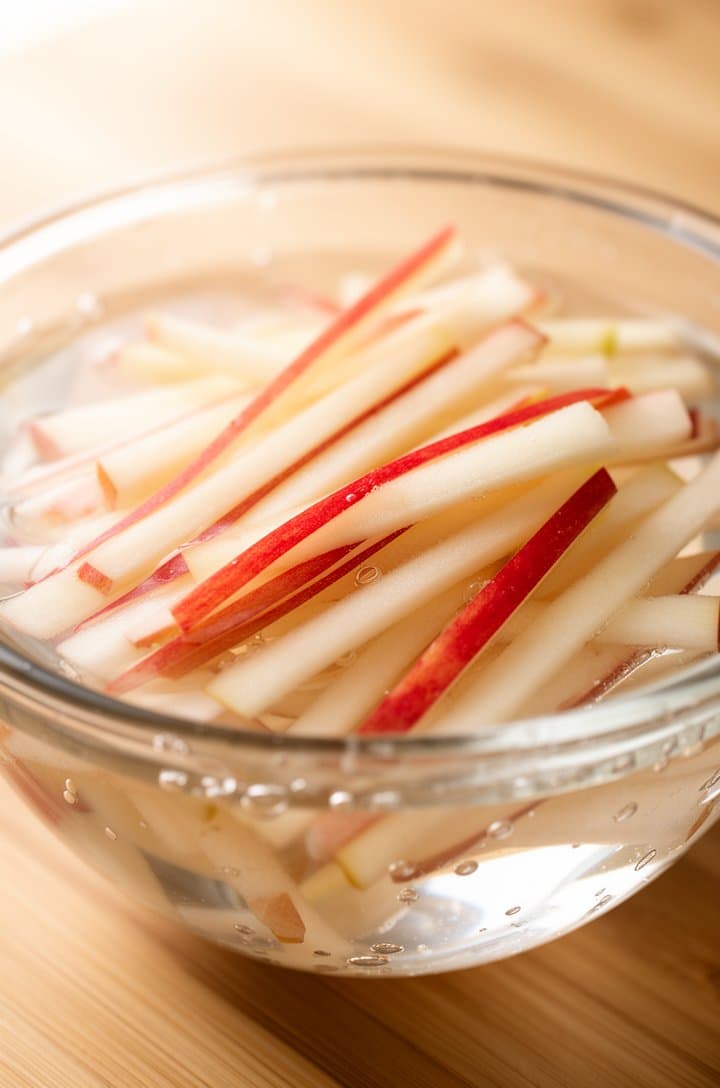 Side-angle close-up of thin apple matchsticks soaking in a clear glass bowl of water, showing the crisp white flesh and streaks of red skin, a few bubbles visible in the water, soft natural backlighting creating a glow through the glass, light wooden surface underneath