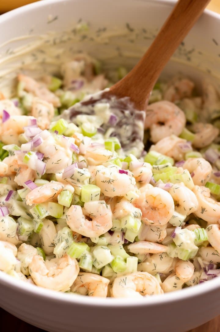 45-degree angle close-up of chopped poached shrimp, diced celery, and finely minced red onion being folded together with the creamy lemon-dill dressing in a large white ceramic mixing bowl, a wooden spoon mid-stir showing the glossy coating on each piece, flecks of green dill visible throughout, warm natural side lighting