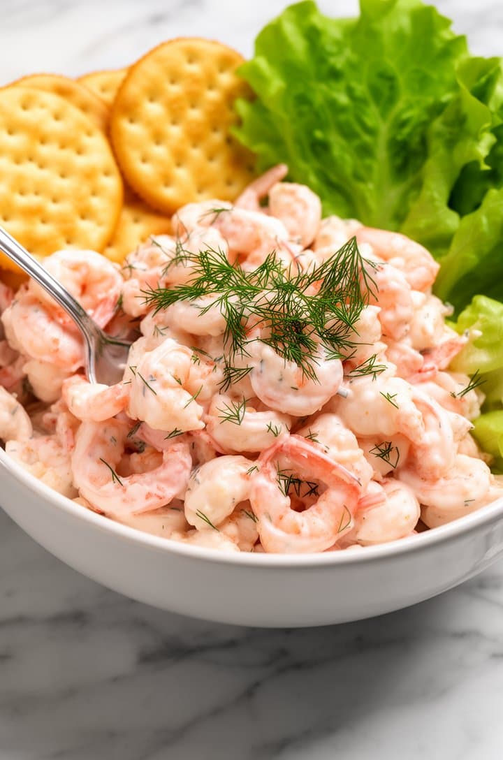 Straight-on close-up of finished shrimp salad served in a shallow white ceramic bowl on a marble surface, a silver spoon resting in the salad, plump pink shrimp pieces glistening with creamy dressing, fresh dill fronds scattered on top as garnish, golden saltine crackers and green butter lettuce leaves arranged alongside the bowl, bright natural light casting soft shadows, professional food blog styling