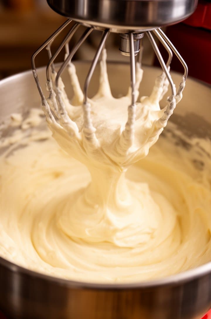 Close-up of the stand mixer bowl showing thick, pale ivory whipped egg-sugar mixture at the ribbon stage, the whisk lifted showing the batter falling in a thick stream that holds its shape, tripled in volume, cloud-like texture, warm kitchen lighting