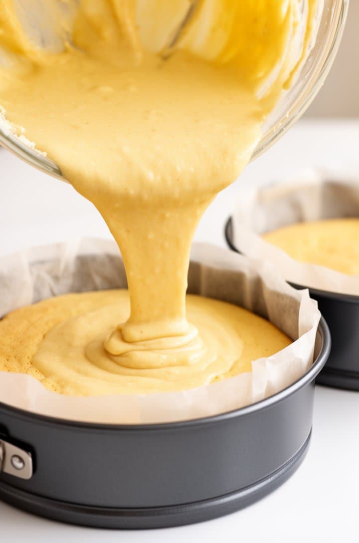 45-degree angle of pale yellow sponge cake batter being poured from the mixing bowl into a parchment-lined 9-inch round cake pan, the batter flowing in a thick smooth stream, second empty pan waiting beside it, bright kitchen lighting, clean white surface