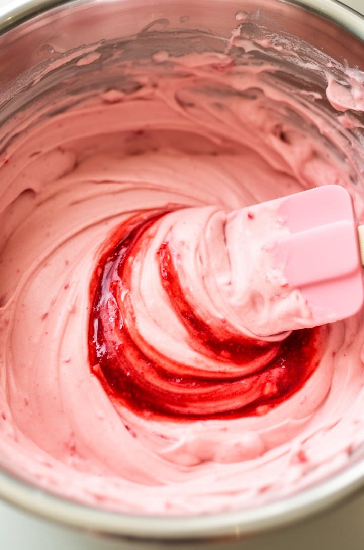 Overhead shot of pink strawberry cake batter in a large stainless steel mixing bowl, smooth and glossy with a beautiful pale pink hue, a rubber spatula folding in the last of the reduced strawberry puree creating marbled streaks, bits of batter on the bowl's rim, bright natural lighting